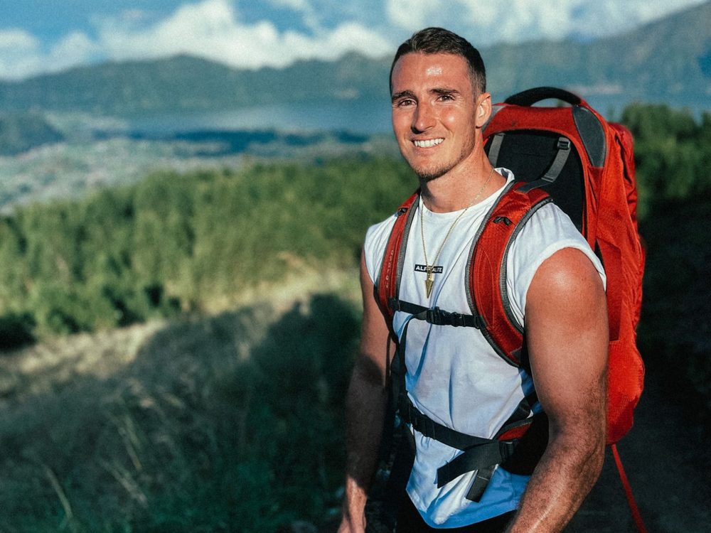Nick Updike smiling on a hike in Bali, red backpack, lake and mountains behind him
